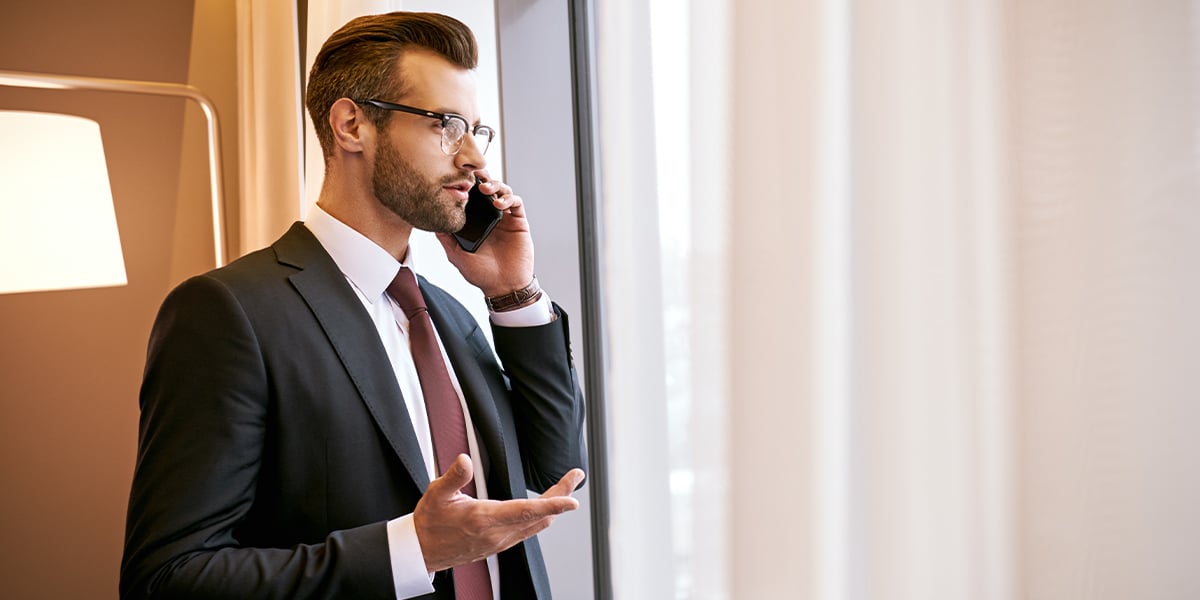 A businessman in a suit standing in front of a floor to ceiling window talking on his smartphone.