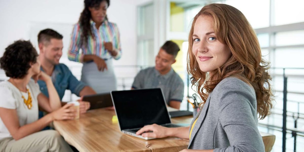 A woman sitting at a conference table with a laptop turning to look at the camera while others in the background are having a conversation while looking at a table.